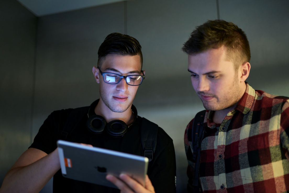 Two young men look at a tablet together, one wearing glasses and headphones, the other in a plaid shirt.